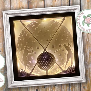 Pode incluir: Uma moldura branca com um passe-partout preto que envolve uma caixa de luz com uma bola de golfe e dois golfistas balançando seus tacos. A caixa de luz é iluminada por trás, criando um efeito de silhueta. A moldura está sobre um fundo de madeira.