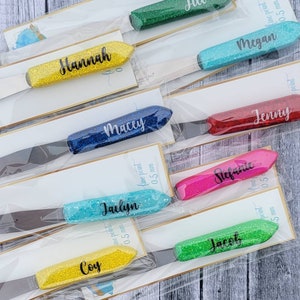 May include: A set of ten personalized glitter paint spatulas with names printed on each. The spatulas are packaged individually in clear plastic bags. The spatulas are in various colors, including yellow, blue, green, pink, red, and turquoise.