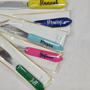 May include: Five personalized butter knives with glitter handles. The handles are yellow, blue, light blue, pink, and green. The names "Hannah", "Macey", "Megan", "Stefanie", and "Jill" are written on the handles.