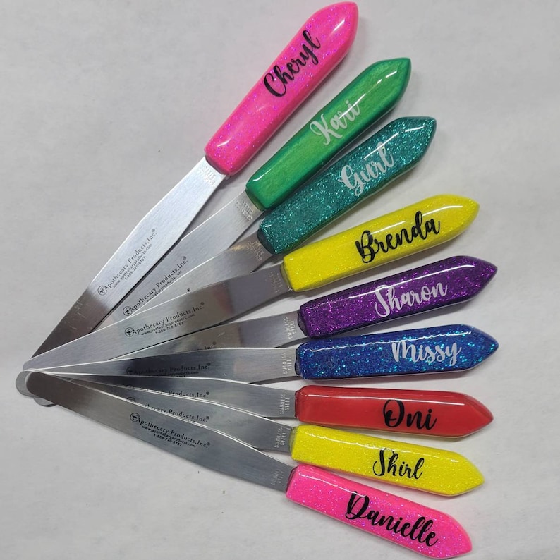 May include: Ten colorful, glittery, personalized spatulas with silver metal blades. Each spatula has a different name on it, including Cheryl, Kari, Gurt, Brenda, Sharon, Missy, Oni, Shirl, and Danielle. The spatulas are made by Apothecary Products, Inc.