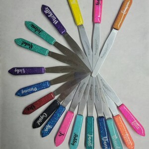 May include: A set of 15 personalized spatulas with glitter handles. Each spatula has a different name on it, including Brandi, Mercury, Dana, Janet, Ashley, Ir, Kim, Crystal, Dee, Lillie, Stelania, Vicky, Rachel, Jolene, and Michelle. The spatulas are arranged in a fan shape, with the handles pointing outwards.