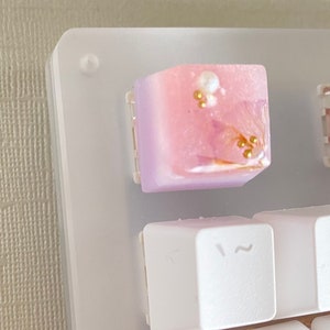 May include: A pink and purple keycap with gold and white accents. The keycap has a gradient design and is decorated with small gold beads and a white pearl.