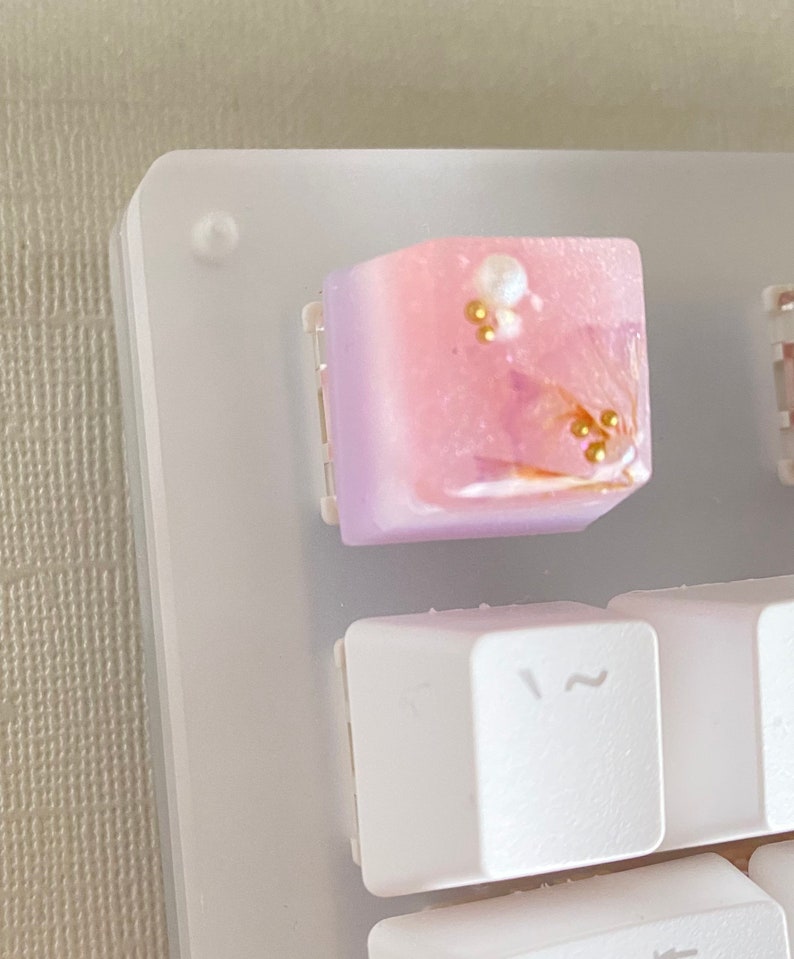 May include: A pink and purple keycap with gold and white accents. The keycap has a gradient design and is decorated with small gold beads and a white pearl.
