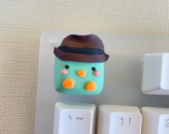May include: A light green and brown clay figurine of a penguin wearing a brown hat. The figurine is sitting on a white keyboard with the keys "Tab", "~", "1!" and "Q" visible.