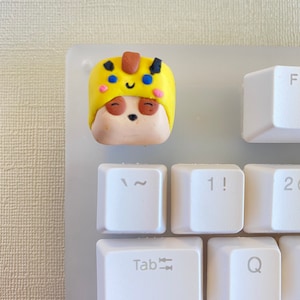 May include: A yellow and brown clay keycap with a cartoon face and a yellow hat. The keycap is on a white keyboard with keys labeled "Tab", "CapsLock", "1!" and "Q".