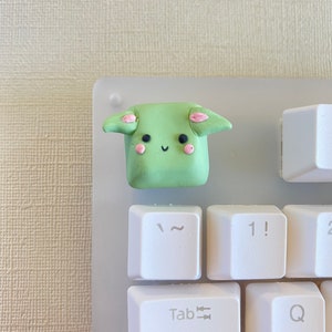May include: A green and pink clay keycap shaped like a cartoon character with ears and a smiling face. The keycap is on a white keyboard.