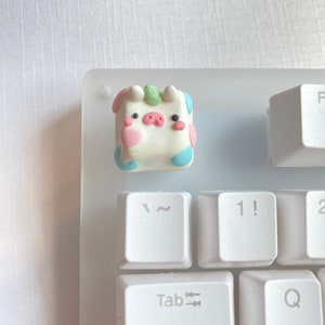 May include: A white, pink, blue, and green cow-shaped keycap with black eyes and a pink nose. The keycap is sitting on a white keyboard.