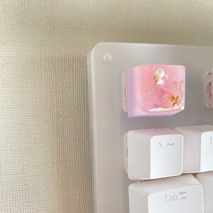 May include: A white keyboard with a pink and white keycap with gold accents and pearl-like embellishments. The keycap is labeled "Tab".