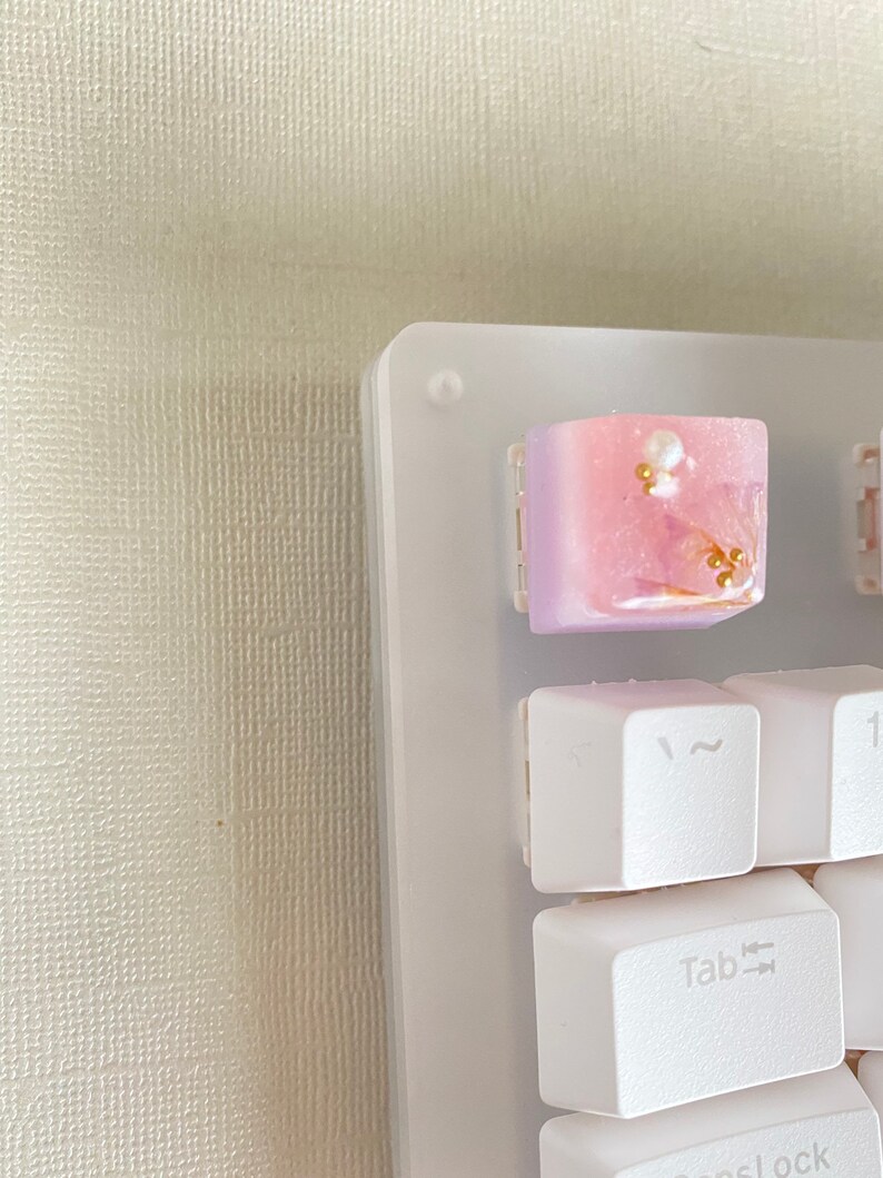 May include: A white keyboard with a pink and white keycap with gold accents and pearl-like embellishments. The keycap is labeled "Tab".