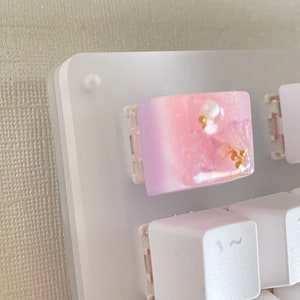 May include: A white keyboard with a pink and purple resin keycap with gold glitter. The keycap is in the top right corner of the keyboard.