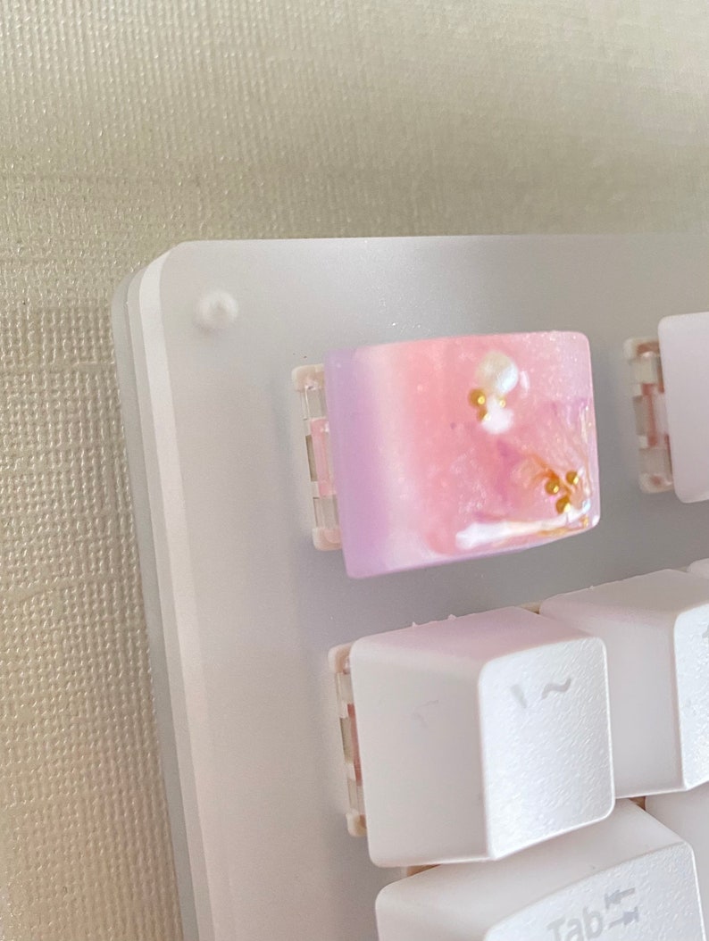 May include: A white keyboard with a pink and purple resin keycap with gold glitter. The keycap is in the top right corner of the keyboard.