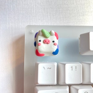 May include: A white keycap with a colorful cow design. The cow has a pink nose, black eyes, and a green leaf on its head. The keycap is on a white keyboard.