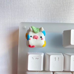 May include: A white, hand-painted, cow-shaped keycap with colorful spots and a green leaf on top. The keycap is sitting on a white keyboard.