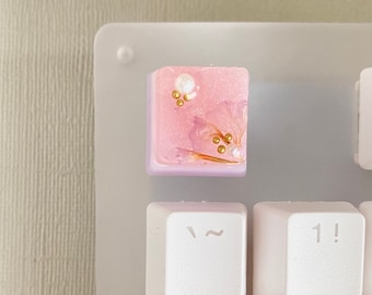 May include: A pink and purple keycap with gold flecks and a white pearl embedded in the resin. The keycap is on a white keyboard.