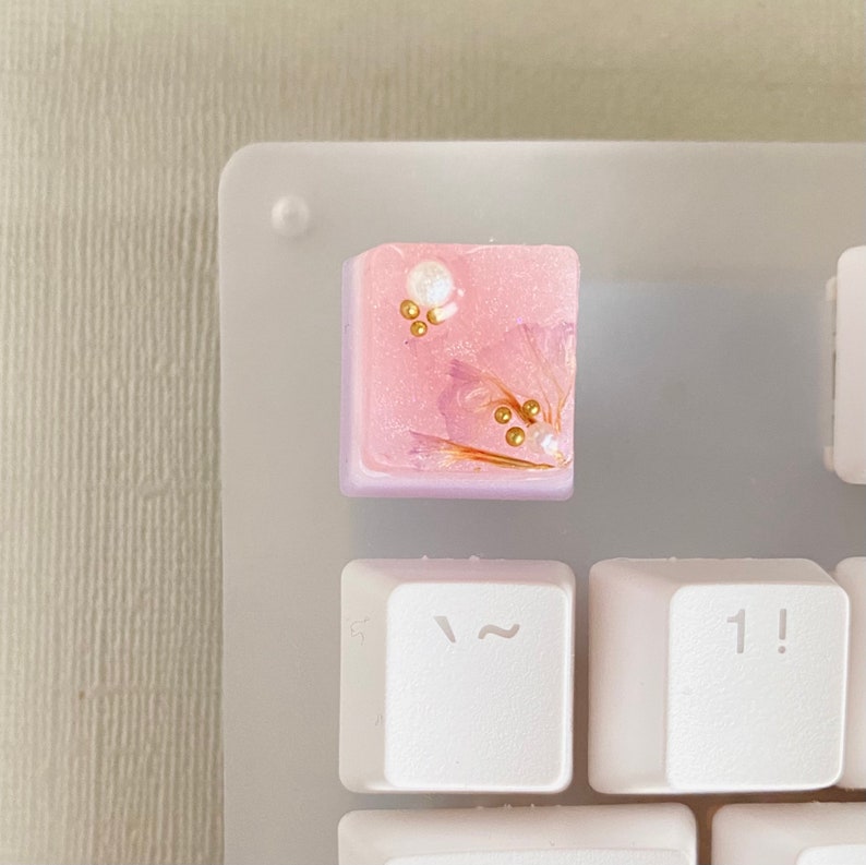 May include: A pink and purple keycap with gold flecks and a white pearl embedded in the resin. The keycap is on a white keyboard.
