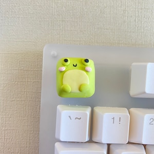May include: A green frog keycap with pink cheeks and black eyes, sitting on a white keyboard with keys labeled "Tab", "~", "1!", "2", and "Q".