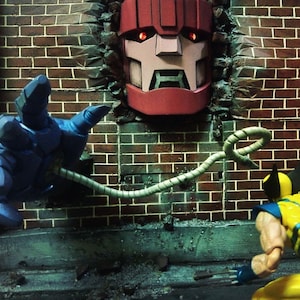 Può includere: Una action figure di robot rosso e blu con un cavo bianco che si estende dal suo braccio, raggiungendo una action figure gialla con un costume blu e giallo. Il robot è parzialmente incassato in un muro di mattoni con il testo "KILL ALL MUTANTS" scritto sul muro.