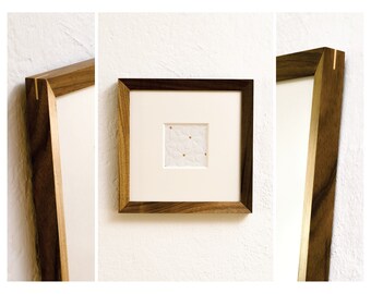 Könnte beinhalten: Ein gerahmtes Kunstwerk mit einem quadratischen, strukturierten weißen Papier mit floralem Muster und goldenen Akzenten. Das Kunstwerk ist in einem dunklen Holzrahmen mit einem goldenen Akzent an der Ecke eingefasst. Der Rahmen ist etwa 25 cm groß.