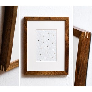 May include: A wooden picture frame with a white mat and a white paper with a pattern of small white flowers and gold dots inside. The frame has a brass inlay on the corners.