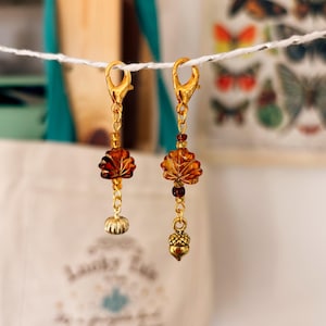 May include: Two gold charm earrings suspended from a white string. Each earring has a gold clasp, amber leaf-shaped beads, and dangling charms. One earring has a small pumpkin charm, and the other has an acorn charm. The background is blurred.