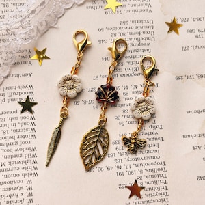 May include: Three decorative charms with gold-colored clasps. One charm features a white flower and a feather, another has a red leaf and a gold leaf, and the third has a white flower and a bee. The charms are on a vintage paper background.