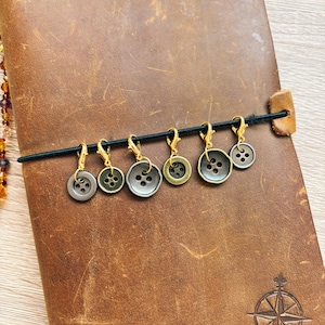 May include: A set of six circular buttons in varying shades of grey and bronze, each attached to a small gold-toned lobster clasp. The buttons feature four holes in the centre and are suspended from a thin black elastic cord.