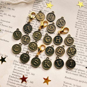 May include: A selection of bronze alphabet charms with gold clasps, displayed on a vintage book page. Each charm displays a different letter. Decorative star-shaped embellishments are also present.