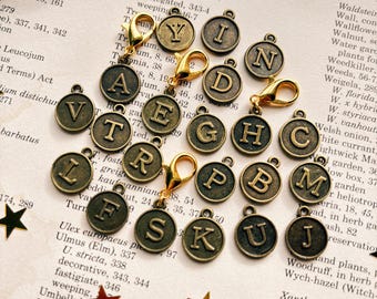 May include: A collection of bronze-colored alphabet charms with gold-colored clasps, scattered on a vintage book page. Each charm features a different letter of the alphabet. Decorative star-shaped embellishments are also present.