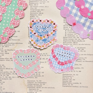 Könnte beinhalten: Drei herzförmige Aufkleber mit Text und dekorativen Rändern. Einer sagt "JOURNAL GIRL" in Blau, ein anderer "STICKER LOVER" in Schwarz und der letzte "I LOVE CRAFTS" in Pink. Der Hintergrund ist eine Vintage-Buchseite.