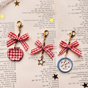 May include: Gold-coloured charm earrings with red and white gingham bows. One earring has a red apple charm, the other a blue and white button design with a cherry illustration. The earrings hang from gold-coloured clasps.