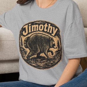 May include: A heather gray short-sleeve t-shirt featuring a circular graphic print of a bear walking through a forest. The text "Jimothy" is arched in a bold, dark font above the bear illustration. The design uses black and tan ink colors.