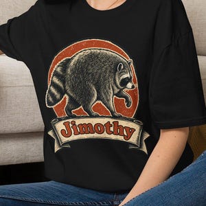 May include: A black short-sleeve t-shirt featuring a graphic of a raccoon walking inside a distressed orange circle. Below the raccoon is a banner with the text "Jimothy" written in a bold, dark red font. The shirt has a crew neckline.