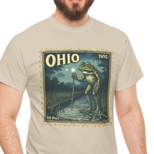 May include: Beige t-shirt featuring a vintage-style graphic of a frog holding a glowing stick in a pond, with the word "OHIO" and the year "1955" in a postage stamp design. The shirt is made of a soft material.