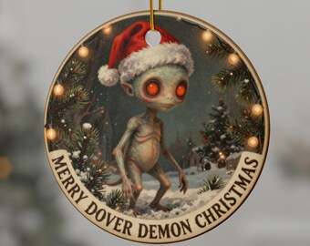 Puede incluir: Adorno navideño redondo de madera con un alienígena gris con un gorro de Papá Noel, ojos rojos y el texto "MERRY DOVER DEMON CHRISTMAS". El adorno cuelga de una cuerda dorada y está rodeado de luces de hadas.