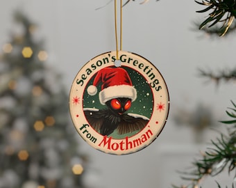 May include: Round Christmas ornament featuring Mothman wearing a Santa hat. The ornament has the text "Season's Greetings from Mothman" in a circular design. The background is green with snowflakes and red stars.