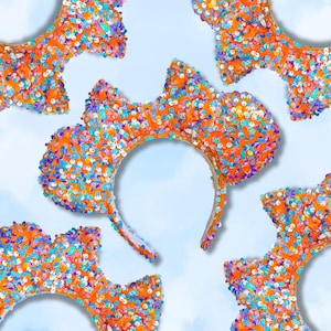 May include: Orange sequined mouse ear headbands with colorful sequins. The headbands have a classic mouse ear shape, with a rounded top and a headband that fits around the head. The ears are covered in orange sequins, and the headband is white.
