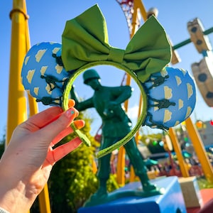 Pode incluir: Orelhas de Minnie Mouse verdes e azuis com um laço verde. As orelhas são decoradas com um padrão de nuvens e pequenas figuras verdes. As orelhas estão sendo seguradas na frente de uma estátua de um soldado de brinquedo verde.