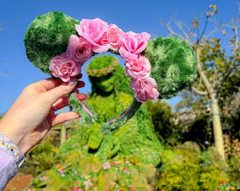 May include: Green fuzzy Minnie Mouse ears with pink fabric flowers. The flowers are arranged in a circle around the headband.