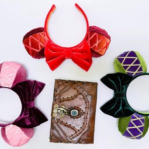 Puede incluir: Tres pares de orejas de ratón de colores con lazos, un par rojo, un par morado y un par verde y morado. Un diario de cuero marrón con un cierre de metal y un diseño de ojo está en el centro.