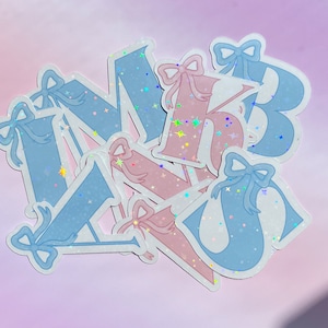 May include: A selection of letter stickers in pastel blue and pink, decorated with bows. The letters feature a holographic, glittery finish, with stars sprinkled across them. The stickers are arranged in a slightly overlapping pattern.