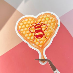 May include: A heart-shaped sticker featuring a honeycomb pattern and a bee in the centre. The sticker is dripping honey and has a glittery finish.