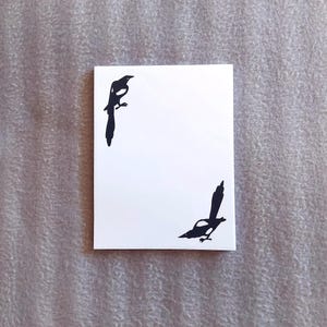 May include: A white card with two black silhouettes of magpies on it.