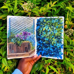 May include: A sketchbook open to two vibrant garden illustrations. One depicts a planter with purple and blue flowers behind a trellis. The other shows a wall of blue flowers and green leaves. The sketchbook is held against a backdrop of green foliage.