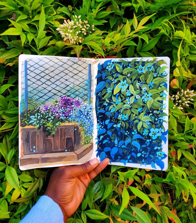 May include: A sketchbook open to two vibrant garden illustrations. One depicts a planter with purple and blue flowers behind a trellis. The other shows a wall of blue flowers and green leaves. The sketchbook is held against a backdrop of green foliage.