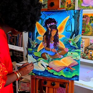 May include: An artist paints a vibrant canvas depicting a fairy with golden wings, surrounded by hummingbirds and a swan. The artwork, set against a blue backdrop, is on an easel. Other paintings are visible in the background, creating an art studio setting.