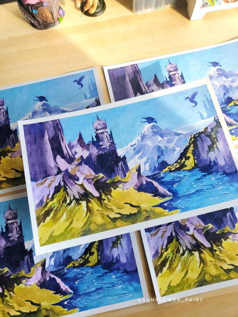 Puede incluir: Una colecci&oacute;n de pinturas de paisajes de fantas&iacute;a con dragones, castillos, monta&ntilde;as y agua. La obra de arte utiliza una paleta de azules, morados, verdes y amarillos. Las pinturas est&aacute;n sobre papel blanco y est&aacute;n dispuestas sobre una superficie de madera clara.