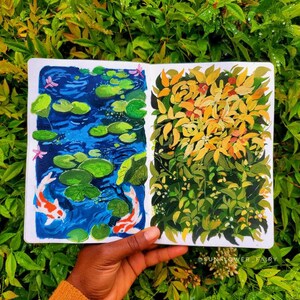 May include: A sketchbook open to two colorful paintings. One depicts a blue pond with lily pads and koi fish. The other shows yellow and green foliage. The sketchbook is held against a backdrop of green leaves.