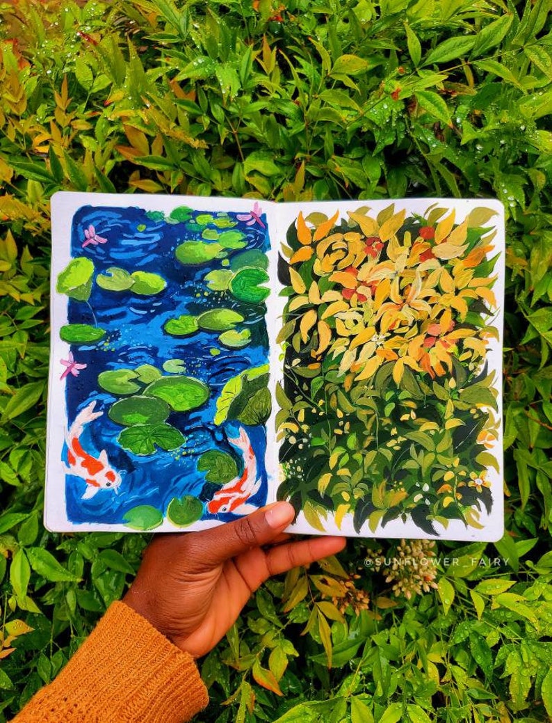 May include: A sketchbook open to two colorful paintings. One depicts a blue pond with lily pads and koi fish. The other shows yellow and green foliage. The sketchbook is held against a backdrop of green leaves.