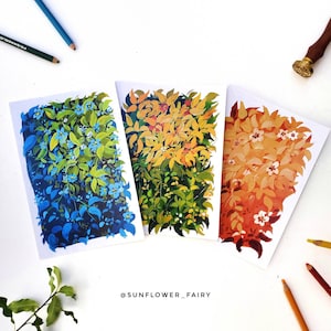 May include: Three botanical art prints featuring vibrant foliage in shades of blue, yellow, and orange. Each print showcases a unique color palette and leaf arrangement. The artist's name, @SUNFLOWER_FAIRY, is visible at the bottom. Colored pencils and a wax seal are also present.