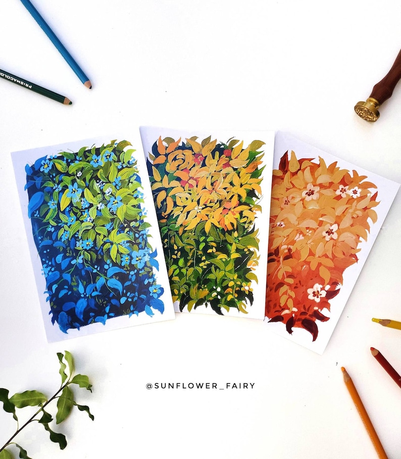 May include: Three botanical art prints featuring vibrant foliage in shades of blue, yellow, and orange. Each print showcases a unique color palette and leaf arrangement. The artist's name, @SUNFLOWER_FAIRY, is visible at the bottom. Colored pencils and a wax seal are also present.